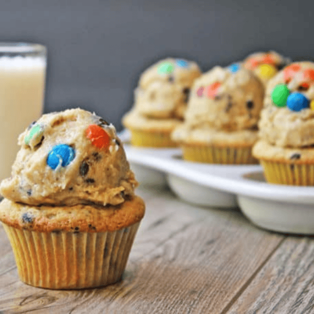 Cake topped with a tan colored icing mixed with candy coated chocolate with a glass of milk and other finished desserts in the background.