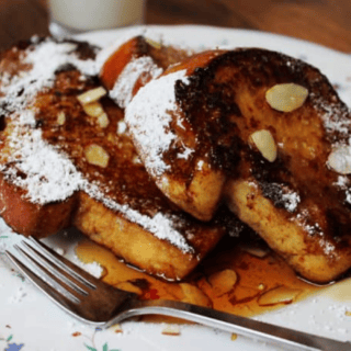Sliced bread that is toasted covered in syrup, powdered sugar and slivers of nuts on a white plate with a fork