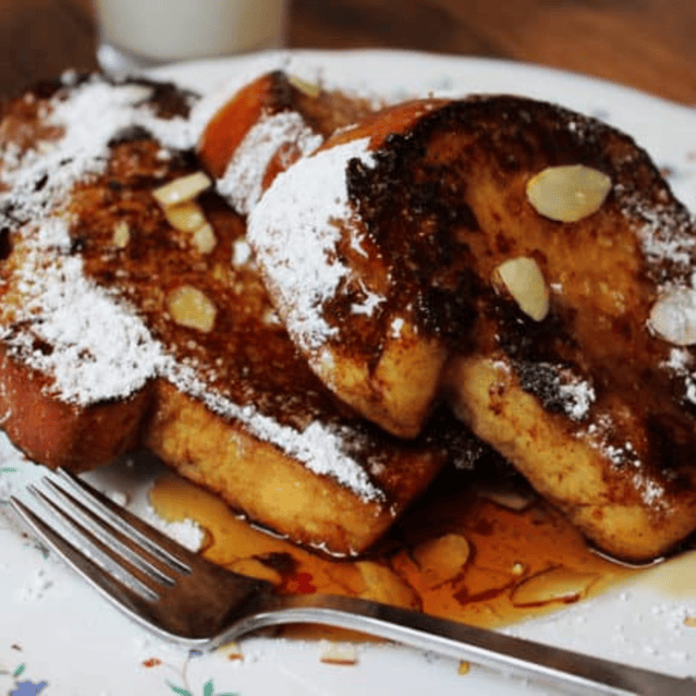 Sliced bread that is toasted covered in syrup, powdered sugar and slivers of nuts on a white plate with a fork