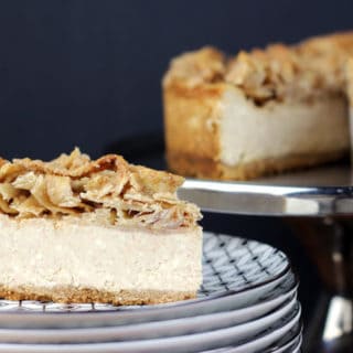 Slice of cheesecake on stack of plates and cake on stand in background.