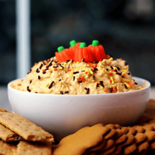 Bowl of pumpkin cheesecake dip with sprinkles and pumpkin candy corn on top, with cookies surrounding
