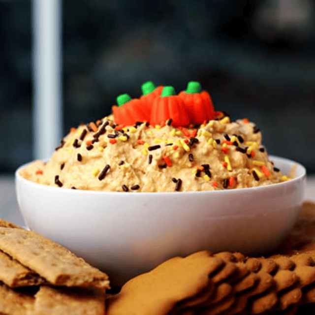 Bowl of pumpkin cheesecake dip with sprinkles and pumpkin candy corn on top, with cookies surrounding