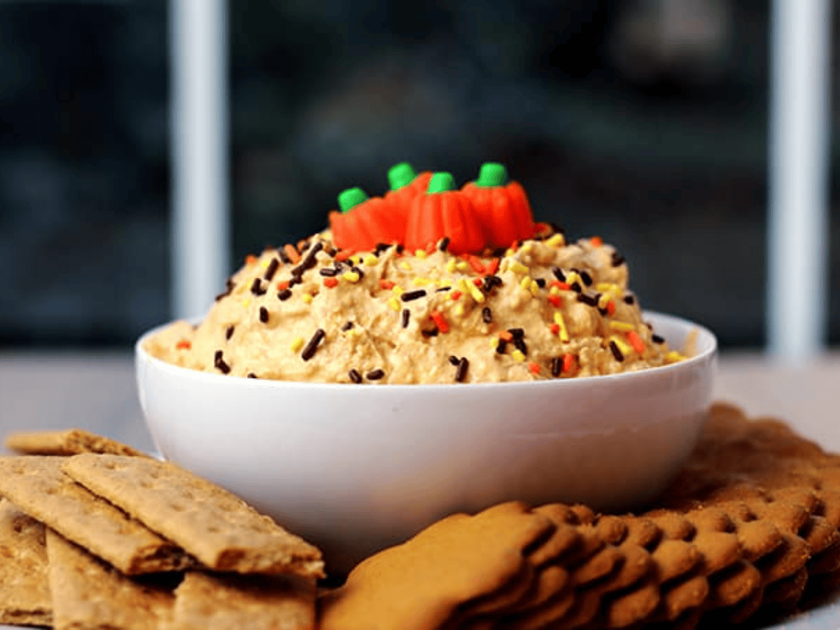 Bowl of pumpkin cheesecake dip with sprinkles and pumpkin candy corn on top, with cookies surrounding