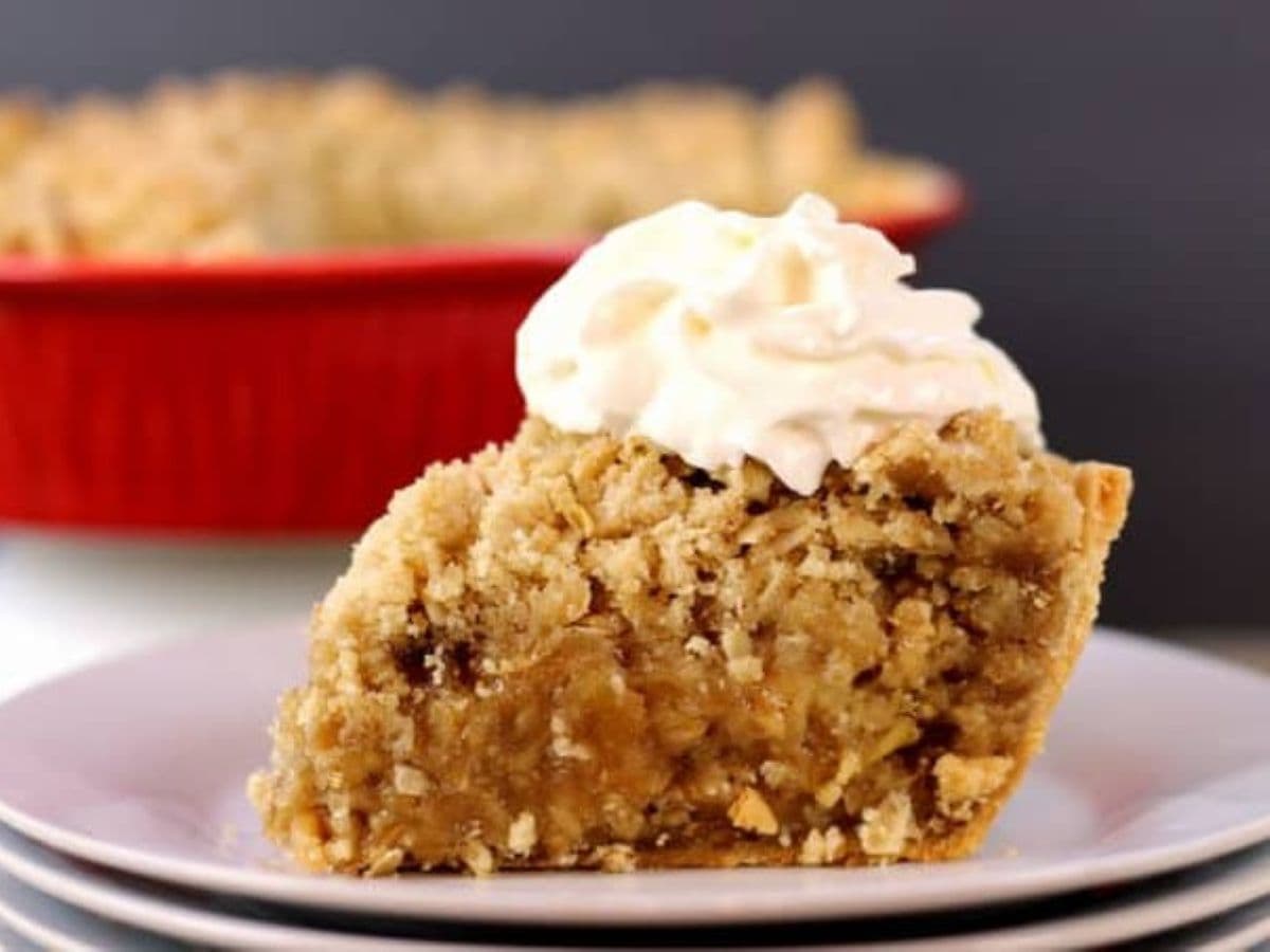 A slice of Apple crisp pie with whipped cream on a stack of white plates.
