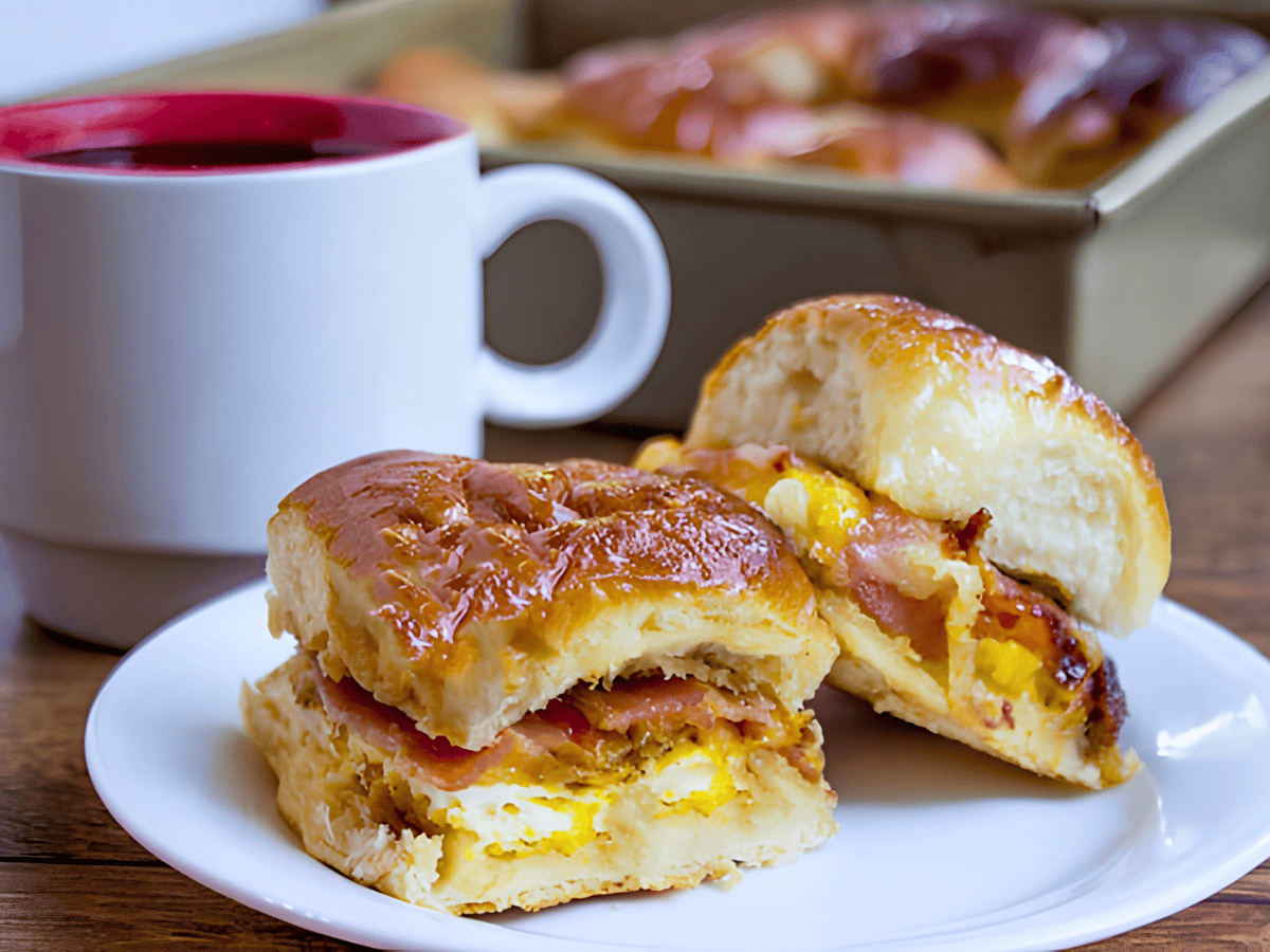 two small breakfast sandwiches on a white plate with a coffee mug on the table