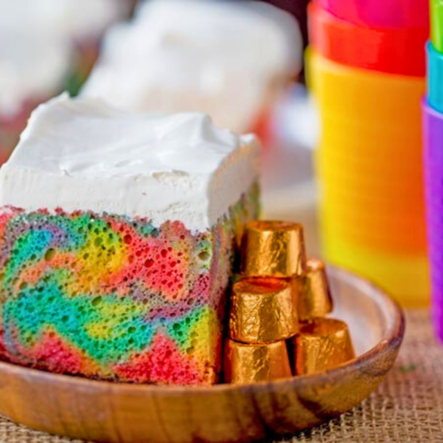 colorful square dessert with frosting on top on a wooden plate with colorful cups on a table
