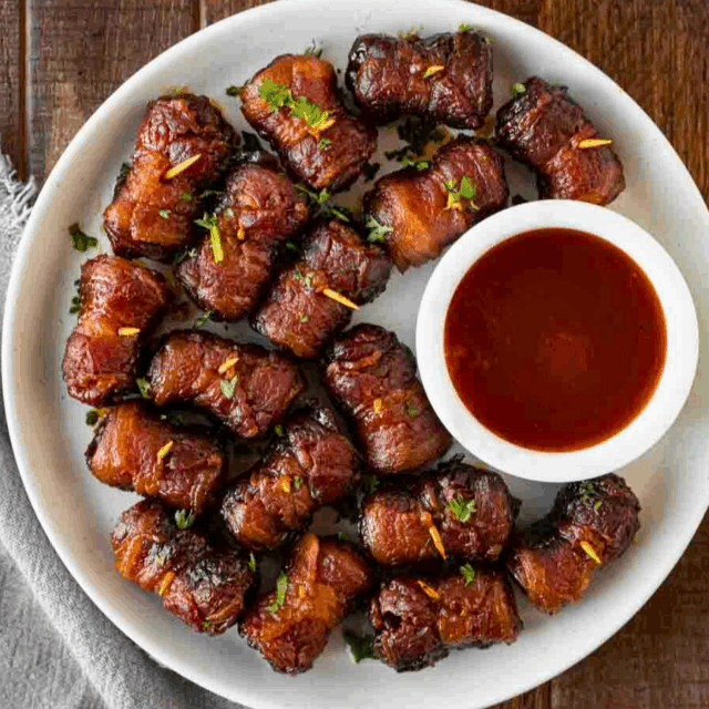 Top down view of cooked bacon wrapped sausages on plate with bowl of BBQ sauce