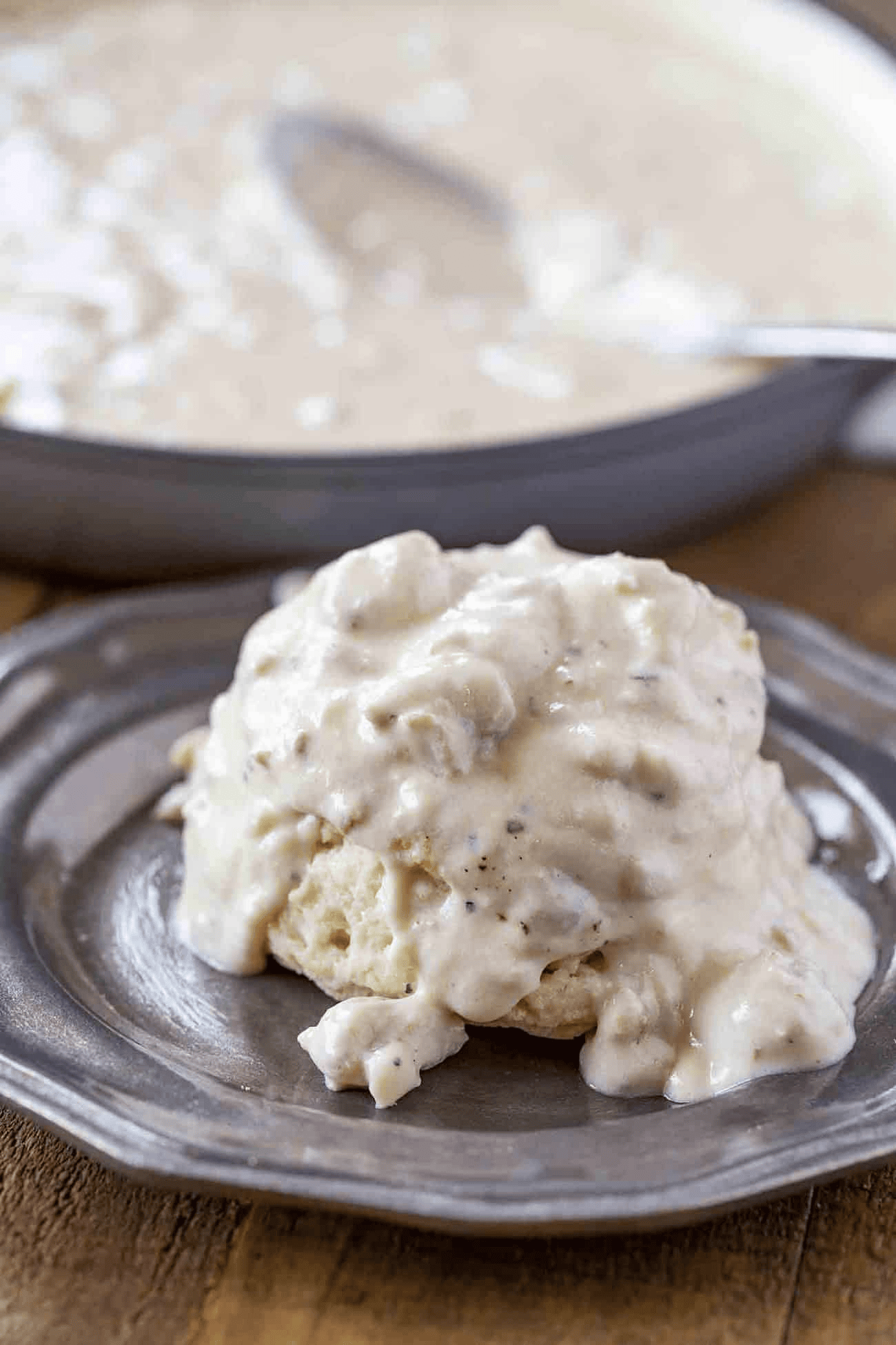 Biscuit on a serving plate smothered in gravy with pan of gravy in the background