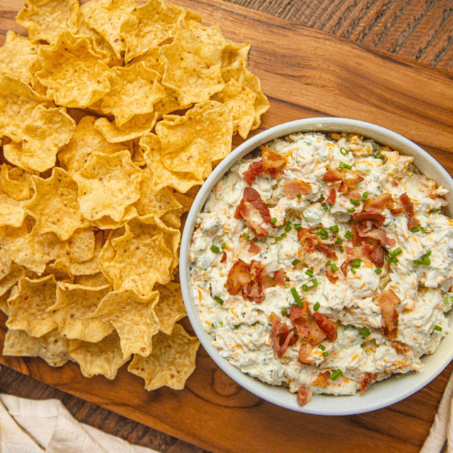 loaded, cheesy, and meaty spread in a bowl next to a pile of chips on a chopping board