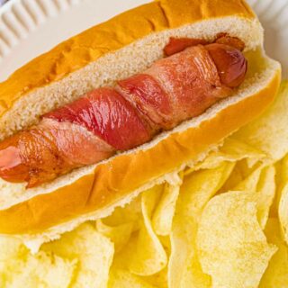 Hot dog wrapped in bacon in a bun with chips