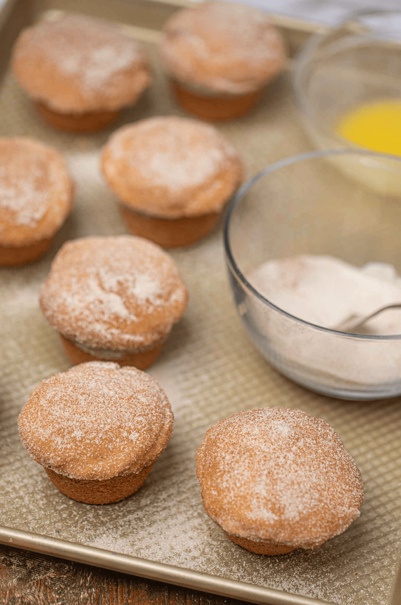 Muffins on a baking sheet with a bowl of cinnamon sugar and a bowl of melted butter to the side