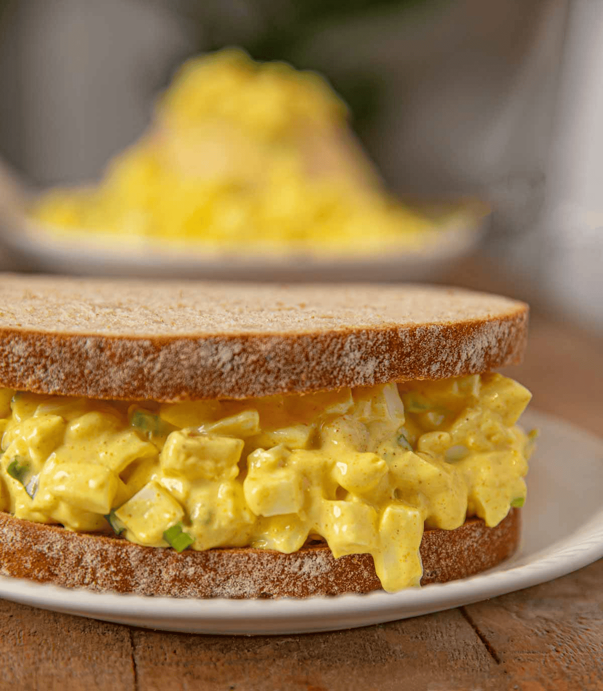 Egg salad sandwich on a plate with bowl of egg salad in the background