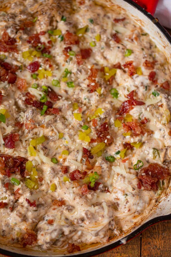 Cheesy Bacon Cheeseburger Dip in red skillet topped with cheese, pickles and sesame seeds