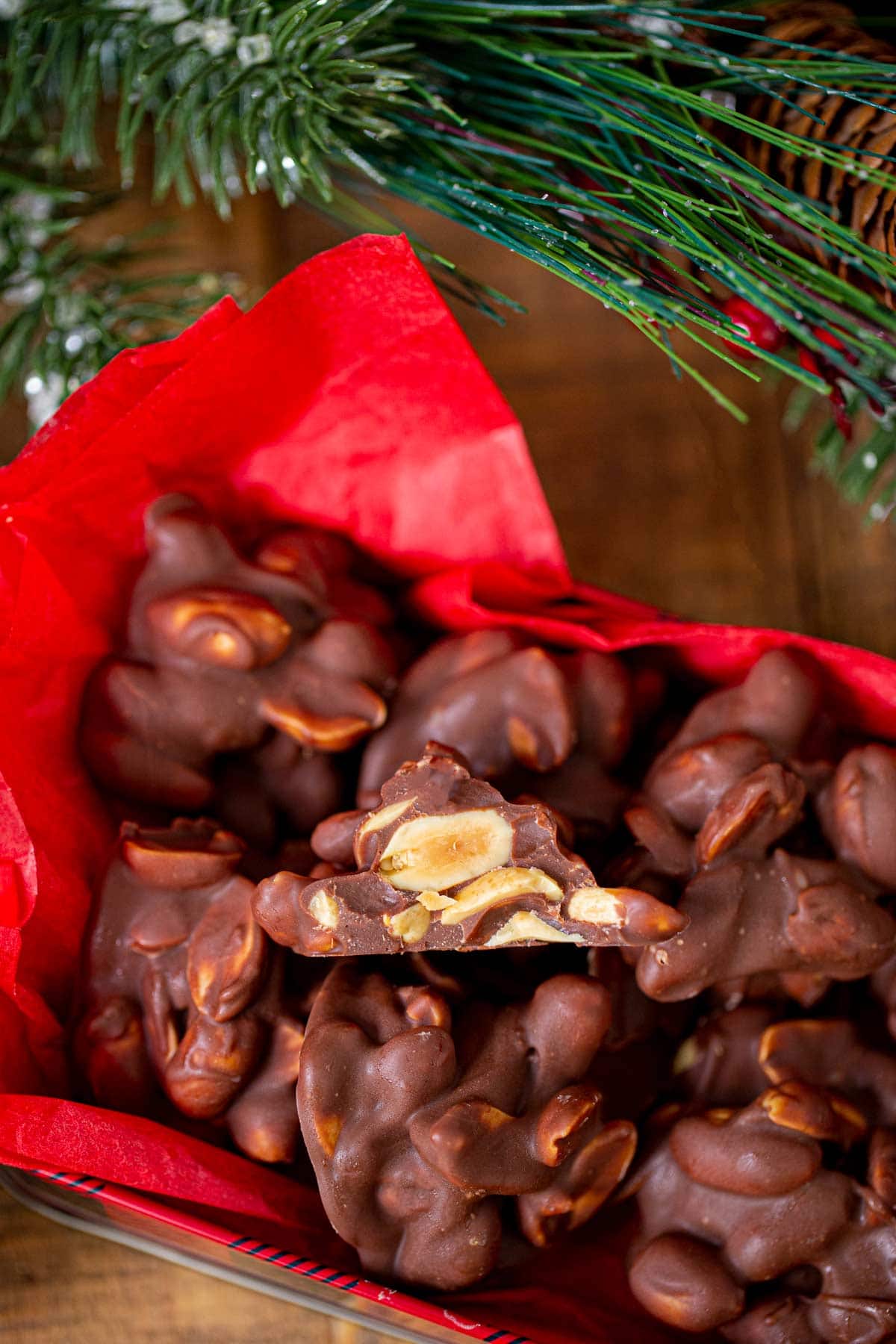 Chocolate Peanut Clusters piled in red tissue lined box with center candy cut in half to show inside