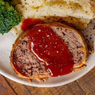 A slice of meatloaf on a white plate with ketchup glaze, mashed potatoes and broccoli,