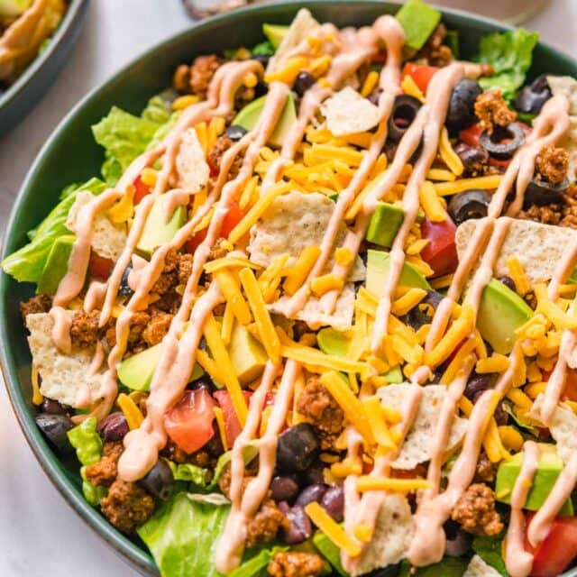 Assembled Taco Salad in bowl with lettuce, black beans, seasoned ground beef, shredded cheese, avocado, and dressing.