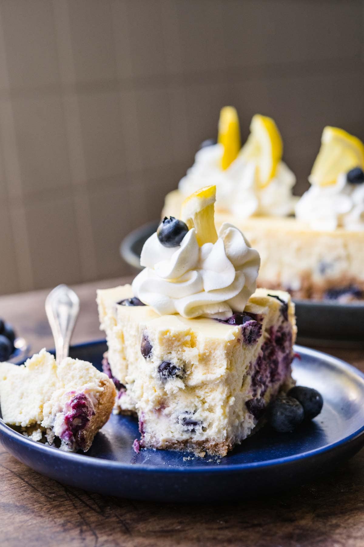 Blueberry Lemon Cheesecake slice on plate with whipped cream, fresh blueberries and lemon garnish