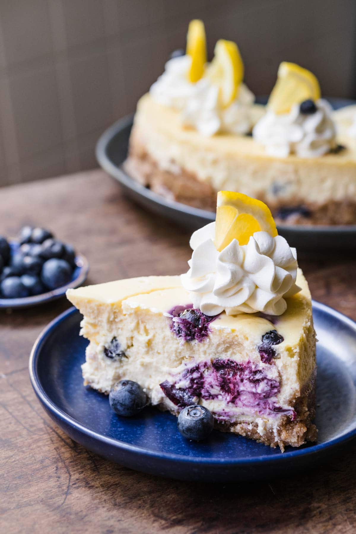 Blueberry Lemon Cheesecake slice on plate with whipped cream, fresh blueberries and lemon garnish