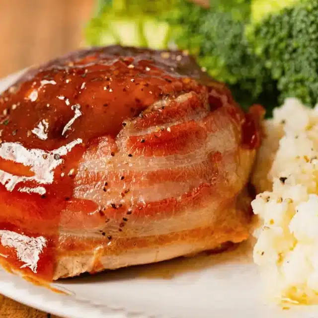 beef roll wrapped in a pork strip on a plate with broccoli and mashed potatoes