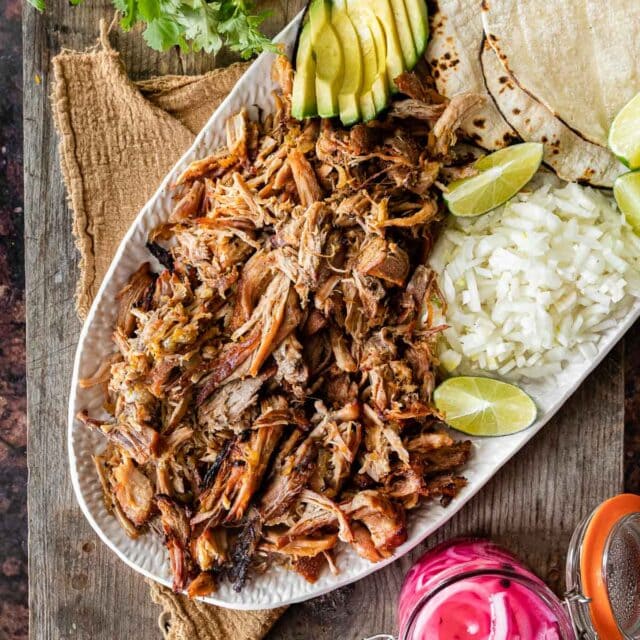 Pork Carnitas (Oven) on serving dish with rice, tortillas, limes, avocado, and pickled onions