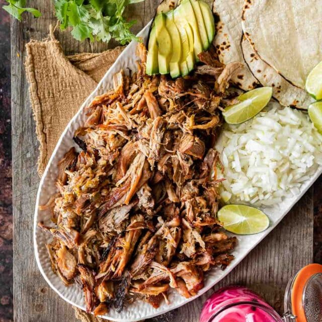 Pork Carnitas (Oven) on serving dish with rice, tortillas, limes, avocado, and pickled onions