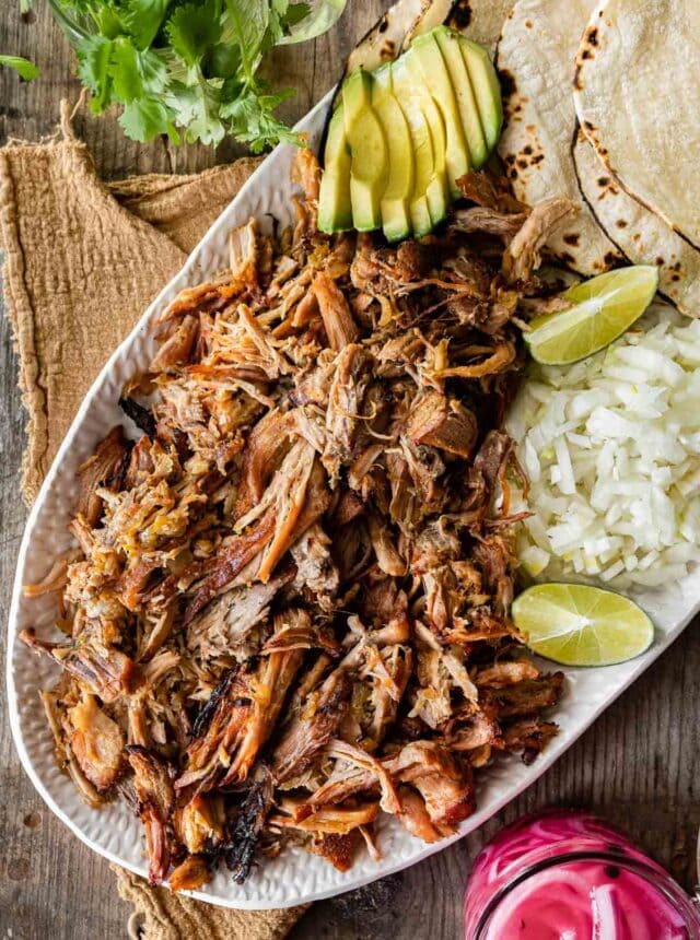 Pork Carnitas (Oven) on serving dish with rice, tortillas, limes, avocado