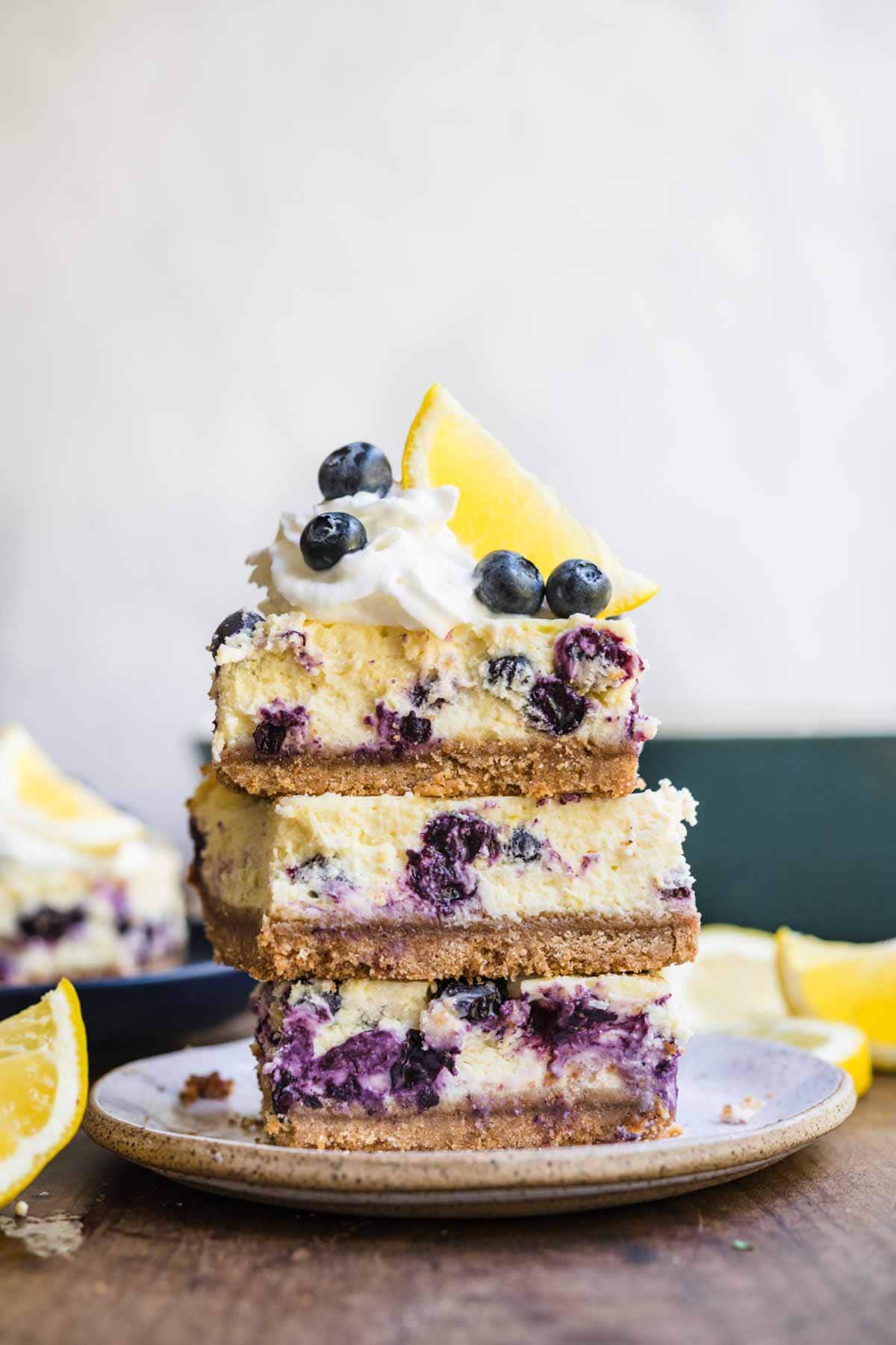 Blueberry Lemon Cheesecake Bars slices stacked on plate garnished with whipped cream, lemon wedge, and fresh blueberries
