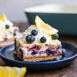 Blueberry Lemon Cheesecake Bars slice on plate garnished with whipped cream, lemon wedge, and fresh blueberries