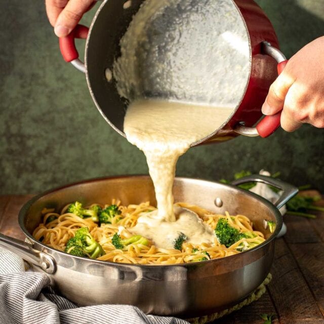 Classic Alfredo Sauce pouring finished sauce over pan of cooked pasta and broccoli. 1x1