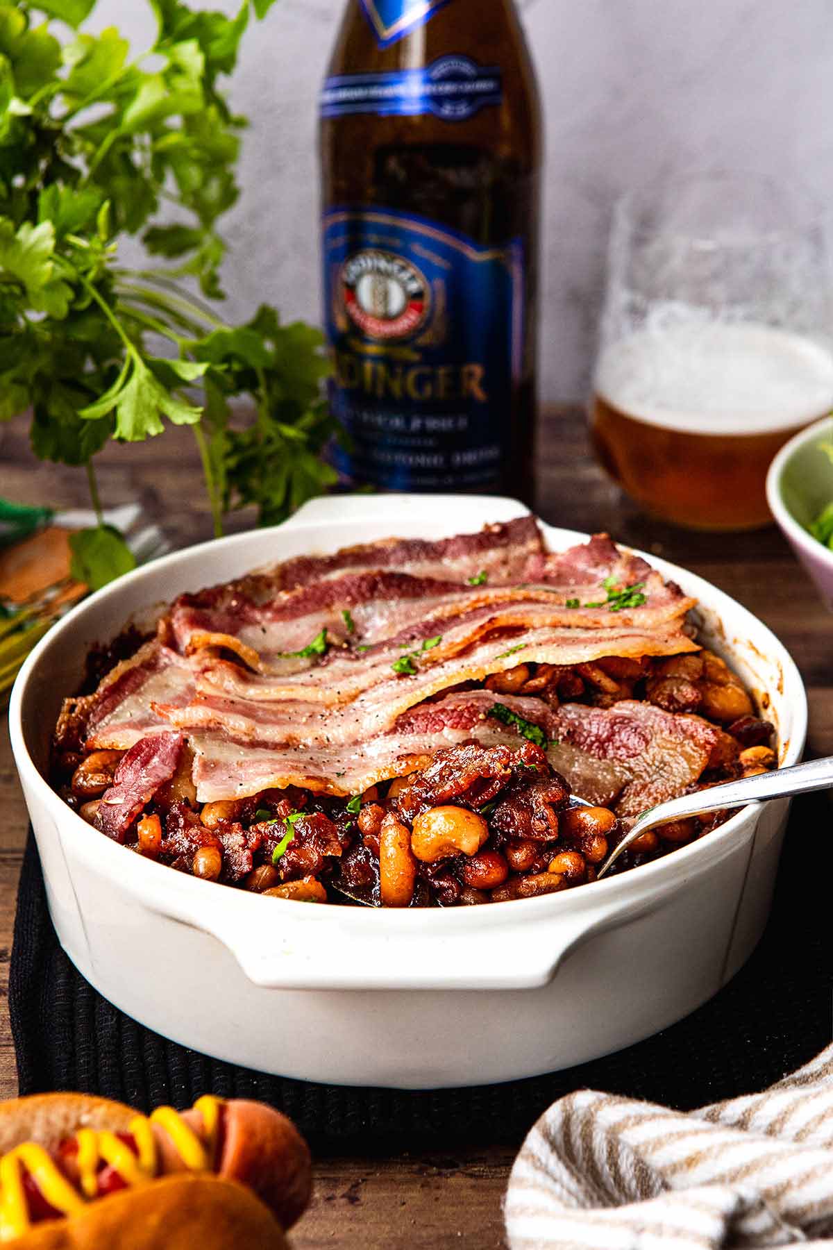 New England Baked Beans in baking dish wtih bacon slices layered on top with serving spoon and beer on the side