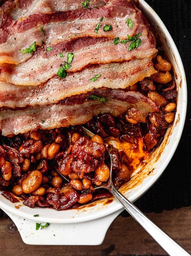 New England Baked Beans in baking dish wtih bacon slices layered on top with serving spoon