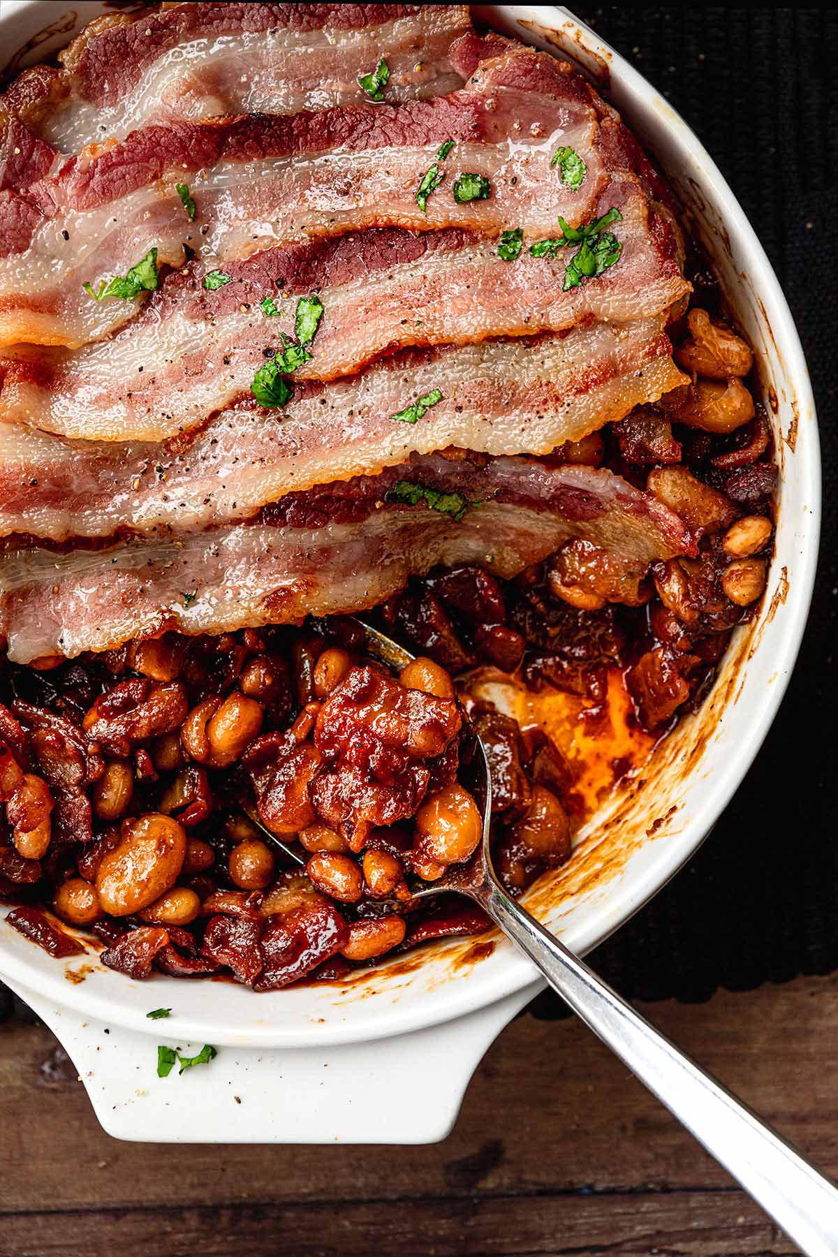 New England Baked Beans in baking dish wtih bacon slices layered on top with serving spoon