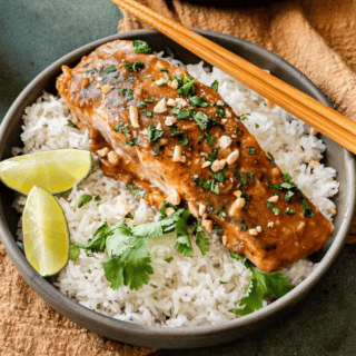 Baked salmon filet with peanut sauce plated over a bowl of rice, garnished with lime wedges.