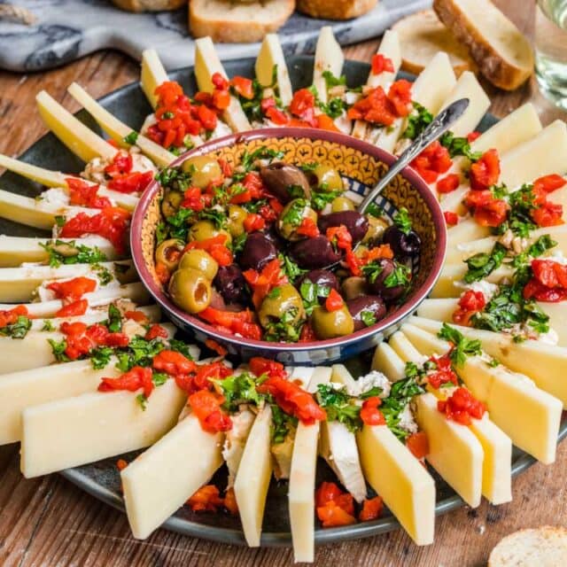 serving platter with olives in center and cheeses arranged around, topped with fresh herbs and pimentos