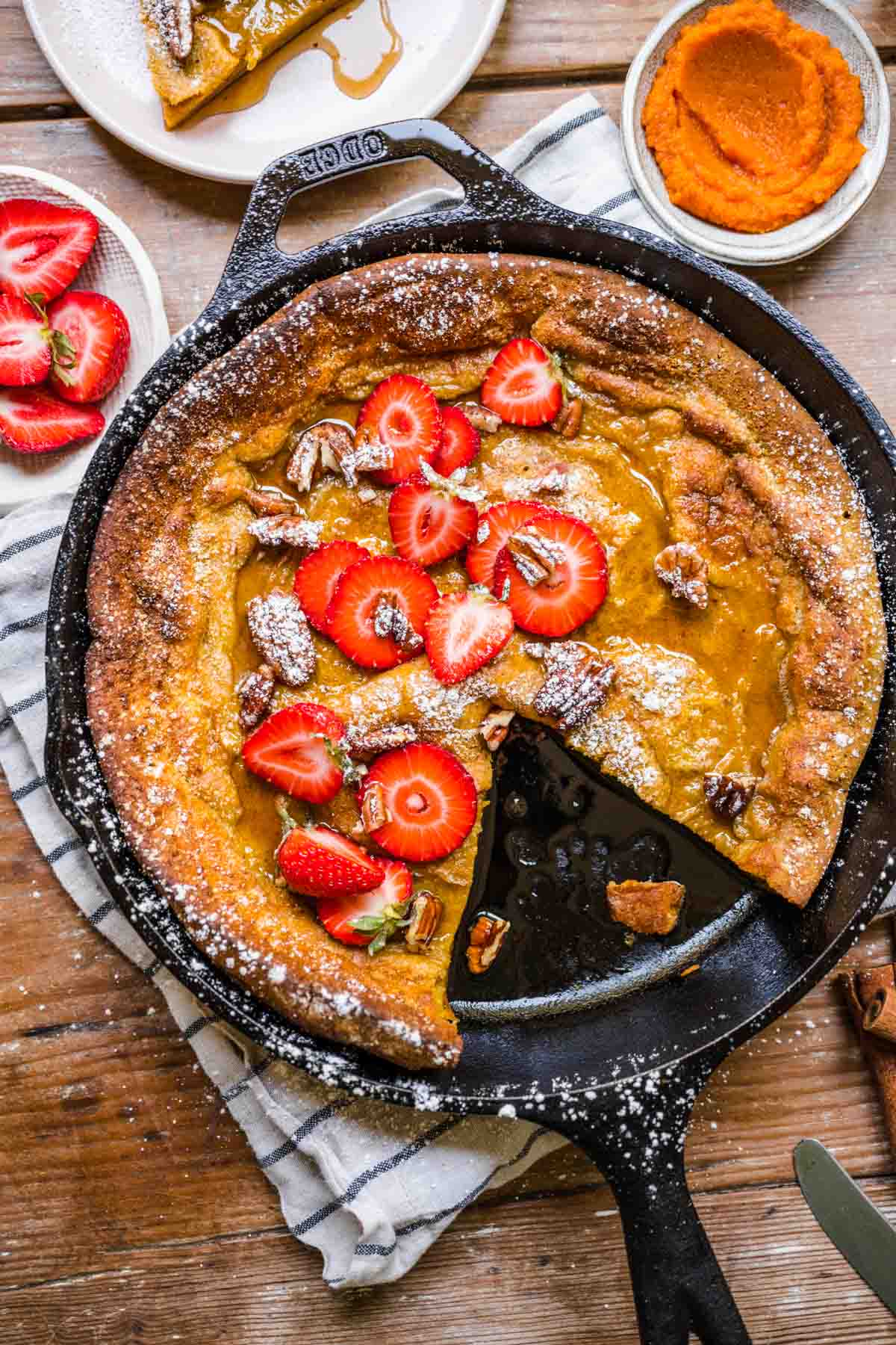 Pumpkin Dutch Baby Pancake baked in skillet with strawberries and powdered sugar on top with slice removed