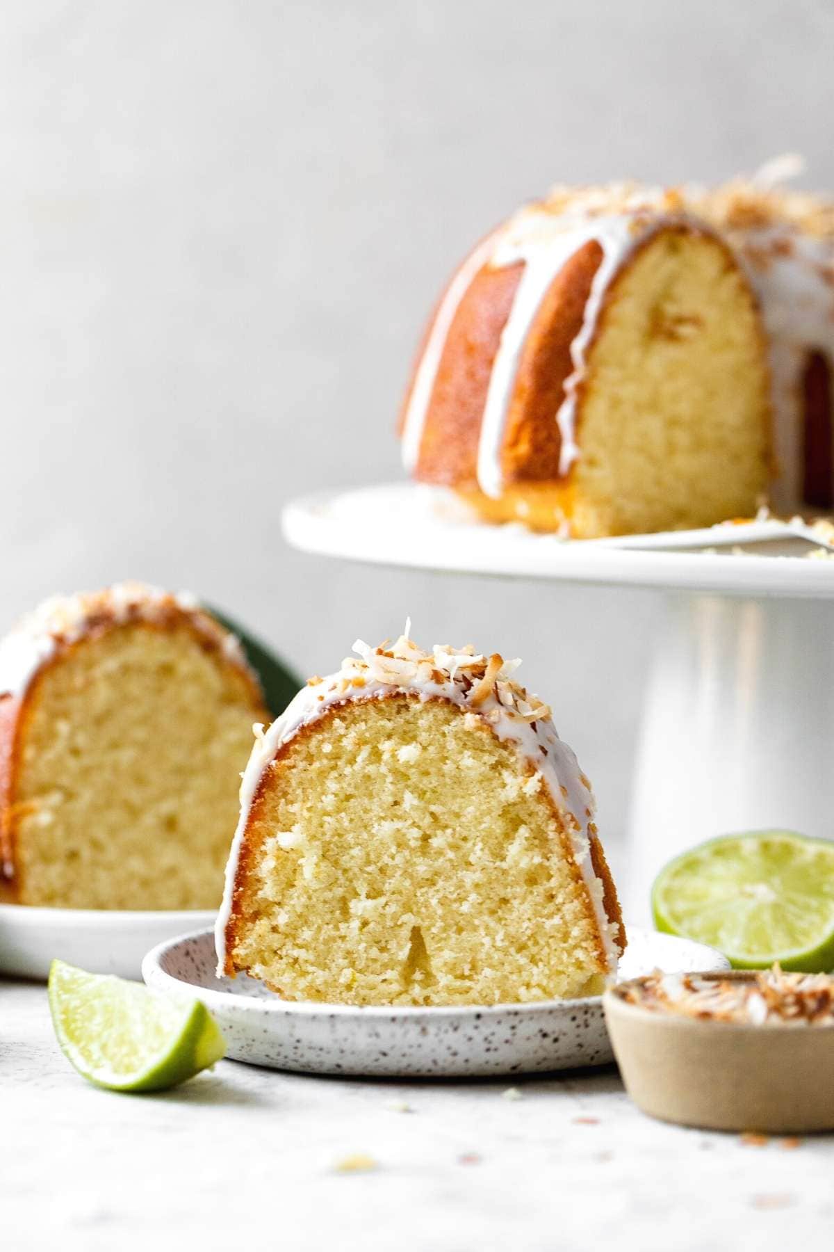Coconut Lime Bundt Cake slice on plate with cake on stand in background