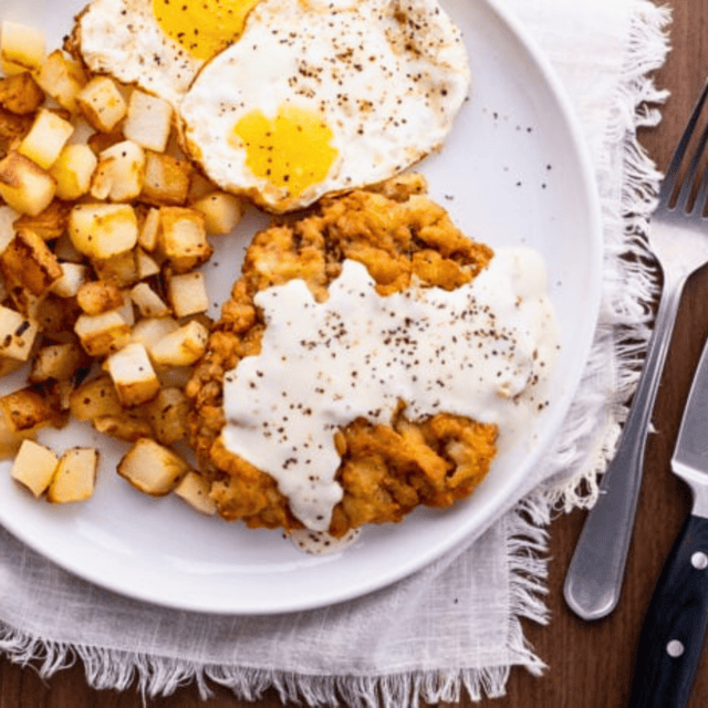 Steak with potatoes and eggs on a plate with silverware next to it