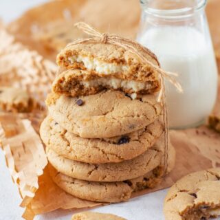 Cheesecake Stuffed Chocolate Chip Cookies baked and stacked on linen towel with top two cut in half to see filling and glass of milk