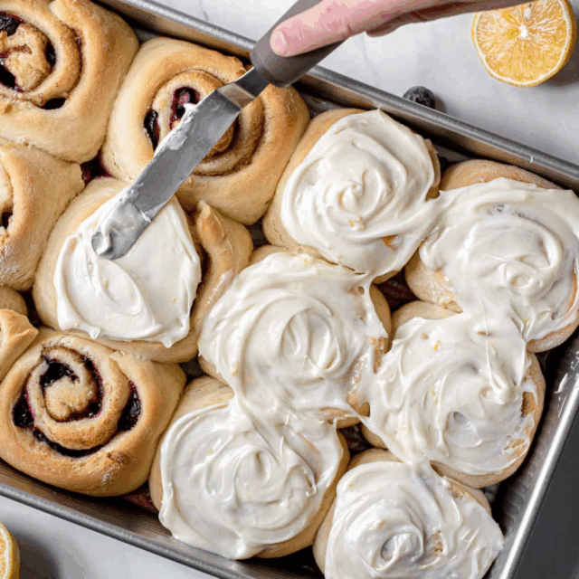 Frosting baked cinnamon rolls in pan with berries and lemon surrounding