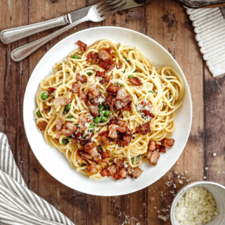Noodles with bacon on top on a white plate with fork and knife next to it