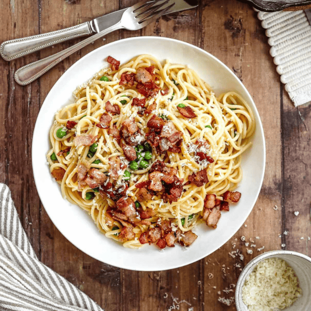 Noodles with bacon on top on a white plate with fork and knife next to it
