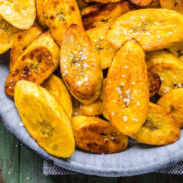 Plantains with bowl of salt next to cooked plantains in blue dish