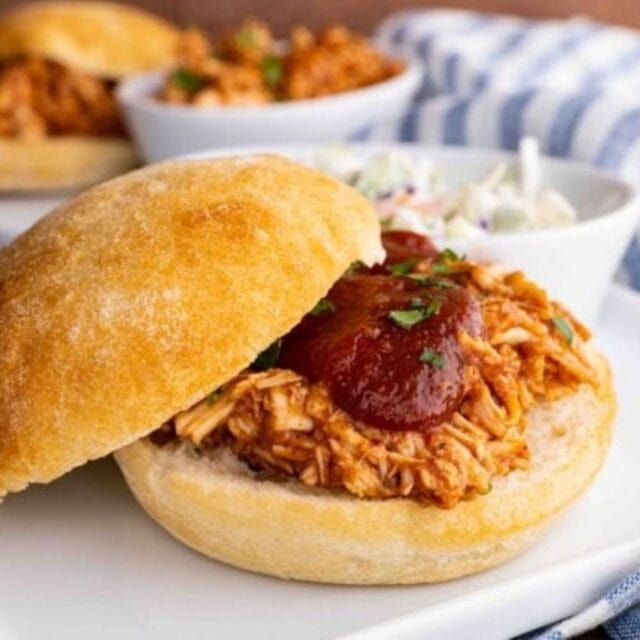 A sandwich on a white plate showing the chicken with a bowl of cole slaw.