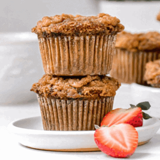 two muffins stacked on white plate with strawberry on the side. more muffins in background