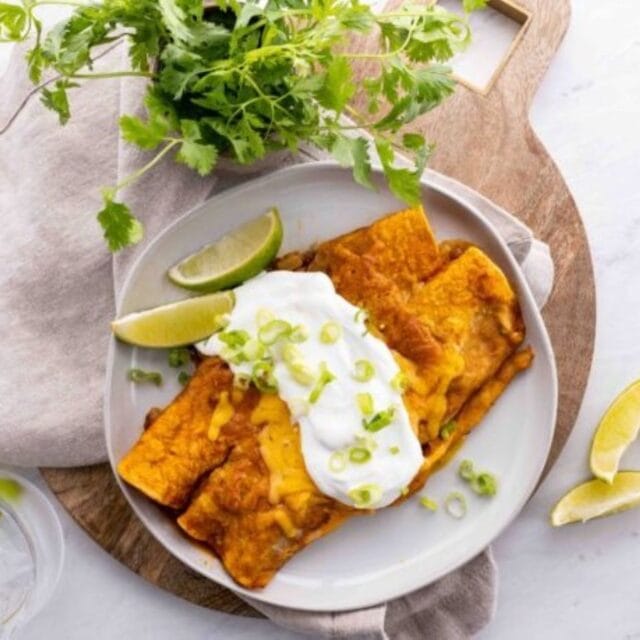 2 enchiladas on a white plate with sour cream and limes.