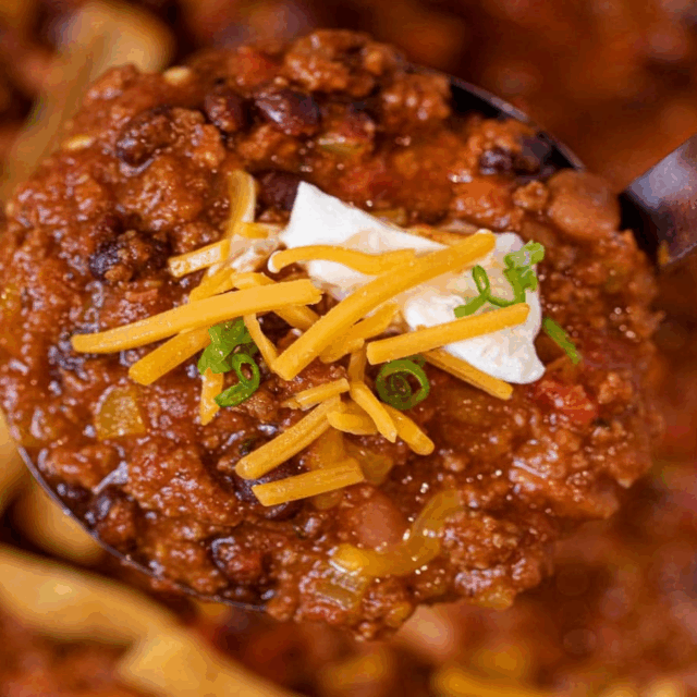 Ladle of chili topped with sour cream, shredded cheese, and scallions.