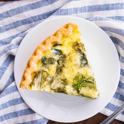 Spinach Quiche slice on plate with tip pointing to bottom right corner. linen napkin with blue stripes around plate.