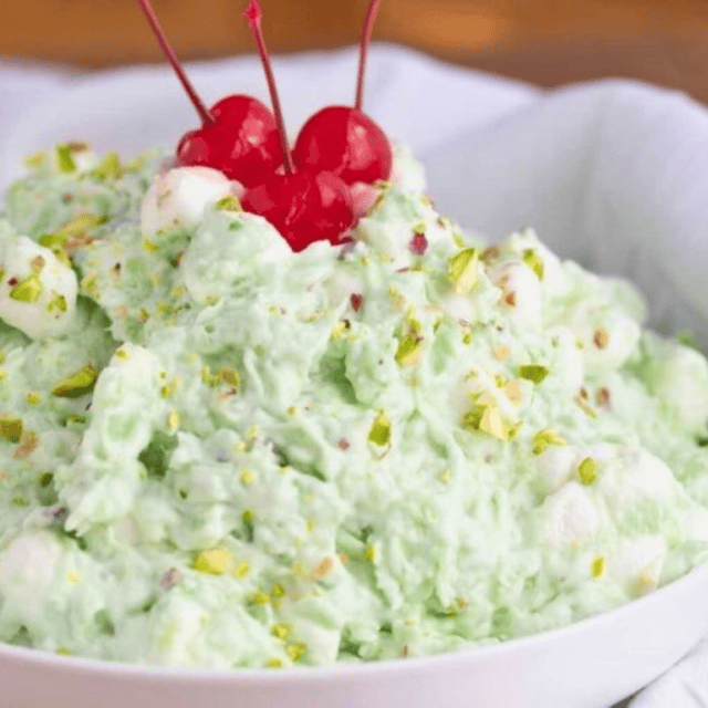 Green fruit salad with marshmallows and chopped pistachios and maraschino cherries on top in white bowl.