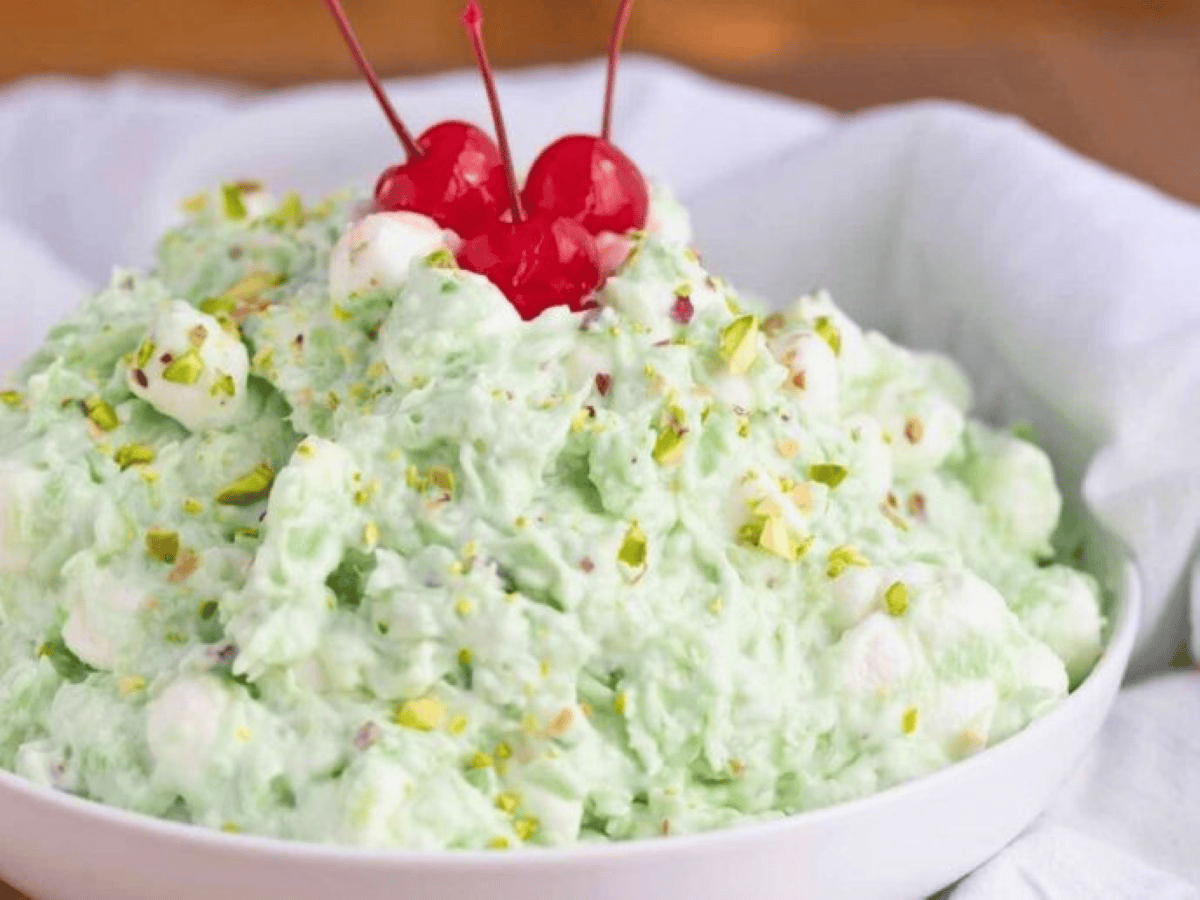 Green fruit salad with marshmallows and chopped pistachios and maraschino cherries on top in white bowl.