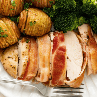 baked chicken wrapped in bacon served with potatoes and broccoli on a white plate with a fork